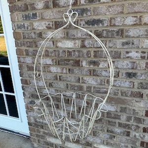 May include: Off-white, hanging metal planter with a decorative oval frame and a basket-like base. The planter is suspended by a hook and is mounted against a weathered brick wall. The design features curved lines and open spaces.
