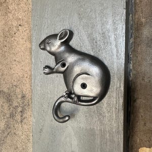 May include: A cast iron mouse-shaped hook in a dark gray finish. The mouse is depicted in a side profile, with a curled tail and two holes for mounting. The hook is mounted on a gray wooden surface.