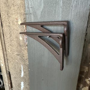 May include: Two brown metal shelf brackets. The brackets are mounted on a weathered gray wooden surface. Each bracket has a triangular shape with a decorative cutout.
