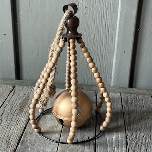 May include: A gold metal bell with a star cutout hangs from a wooden bead and rope garland. The garland is suspended from a metal ring.