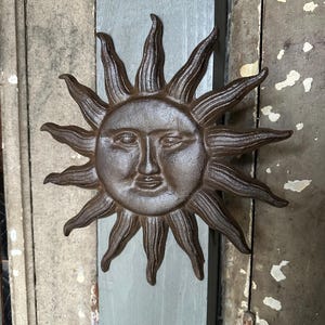 May include: A cast iron sun face wall hanging with radiating rays. The sun has a detailed face with closed eyes and a slight smile. The metal has a rustic, brown finish. The sun is mounted on a weathered, light blue wooden plank.