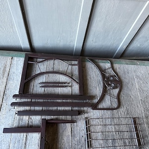Op de afbeelding: Onderdelen van een gedemonteerde donkerbruine metalen decoratieve plantenstandaard. De onderdelen omvatten een rechthoekig frame, gebogen decoratieve stukken en een draadgaasplank. De stukken zijn gerangschikt op een verweerd houten oppervlak.