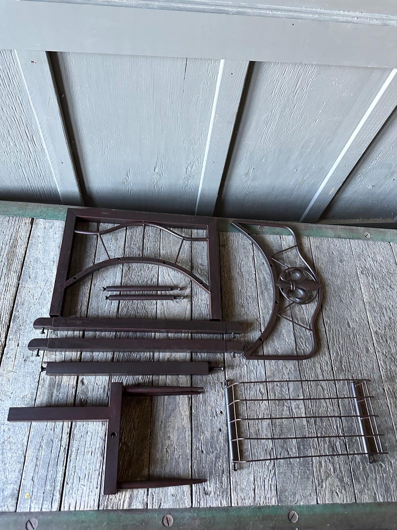Op de afbeelding: Onderdelen van een gedemonteerde donkerbruine metalen decoratieve plantenstandaard. De onderdelen omvatten een rechthoekig frame, gebogen decoratieve stukken en een draadgaasplank. De stukken zijn gerangschikt op een verweerd houten oppervlak.