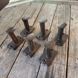 May include: Six rustic brown metal hooks with a rectangular base. The hooks are arranged in two rows of three on a wooden surface.