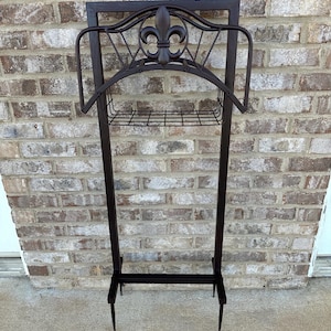 Op de afbeelding: Een donkerbruine metalen tuinslanghouder met een fleur-de-lis-ontwerp. De houder heeft een rechthoekig frame, een gebogen bovenkant en een draadmand voor opslag. De poten zijn puntig.