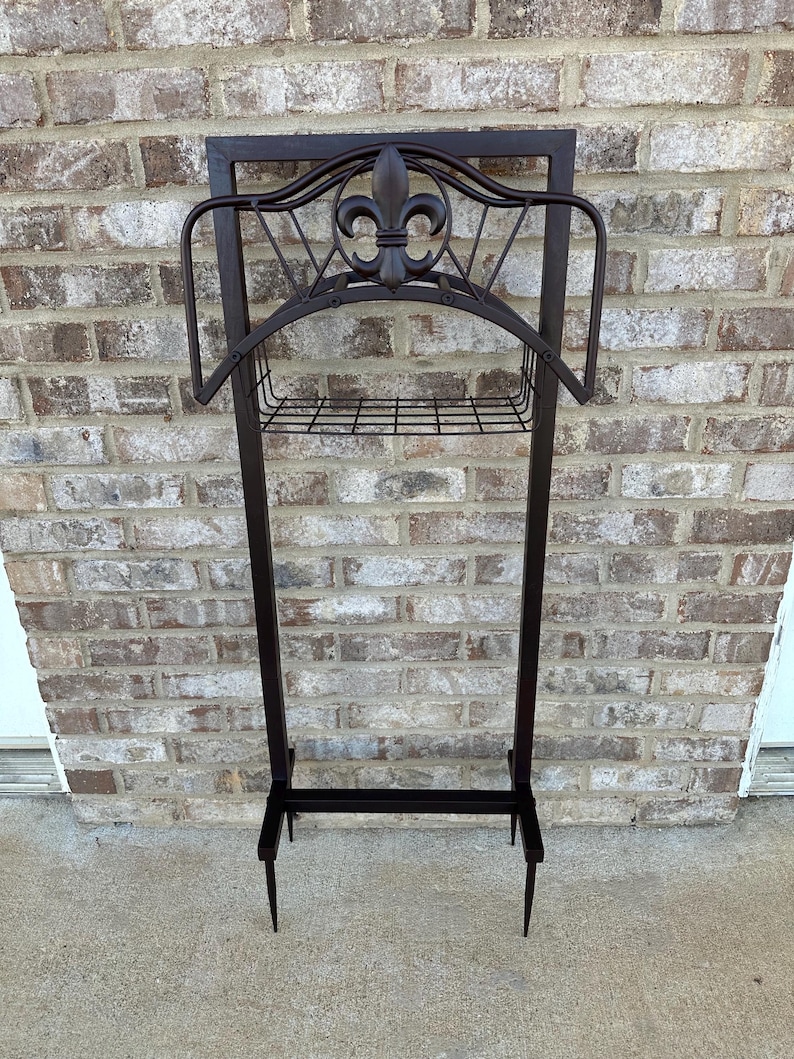 Op de afbeelding: Een donkerbruine metalen tuinslanghouder met een fleur-de-lis-ontwerp. De houder heeft een rechthoekig frame, een gebogen bovenkant en een draadmand voor opslag. De poten zijn puntig.