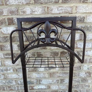 Op de afbeelding: Een donkerbruine metalen tuinslanghouder met een fleur-de-lis-ontwerp. De houder heeft een rechthoekig frame, een draadmand en gebogen armen om de slang vast te houden. De achtergrond is een bakstenen muur.
