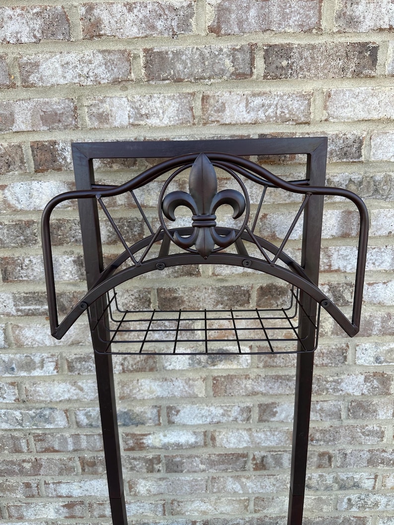 Op de afbeelding: Een donkerbruine metalen tuinslanghouder met een fleur-de-lis-ontwerp. De houder heeft een rechthoekig frame, een draadmand en gebogen armen om de slang vast te houden. De achtergrond is een bakstenen muur.