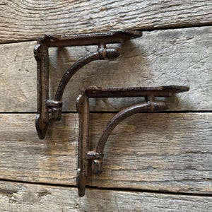May include: Two cast iron shelf brackets with a rustic, aged appearance. The brackets are dark brown with a curved support arm and a flat shelf surface. The design features decorative detailing.