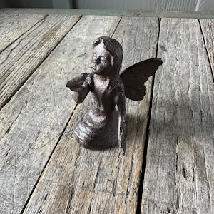 May include: A dark brown cast iron figurine of a kneeling figure with wings, holding a small bird in one hand. The figure has a textured surface and is depicted in a seated position with wings extended behind the back and a bird perched on the hand.