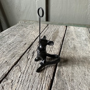 May include: Black metal mouse figurine with a ring on top, used as a photo holder. The mouse is sitting on a wooden surface.