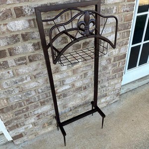 Op de afbeelding: Een donkerbruine metalen tuinslanghouder met een fleur-de-lis-ontwerp. De houder heeft een rechthoekig frame, een gebogen bovenkant en een draadmand voor opslag. Hij staat op twee poten.