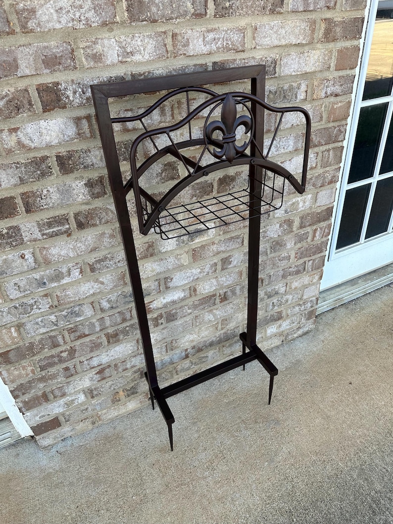 Op de afbeelding: Een donkerbruine metalen tuinslanghouder met een fleur-de-lis-ontwerp. De houder heeft een rechthoekig frame, een gebogen bovenkant en een draadmand voor opslag. Hij staat op twee poten.