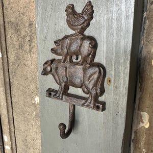 May include: A cast iron wall hook featuring a cow, pig, and chicken stacked on top of each other. The hook is dark brown and mounted on a weathered gray wooden board. The farm animal design adds a rustic touch.