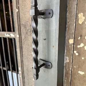 May include: A black, twisted metal door handle with a square base and mounting plates. The handle is mounted on a gray wooden door frame. The door frame is weathered, with visible paint chipping.