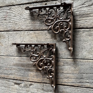 May include: Two ornate, cast iron shelf brackets with a rust-colored finish. The brackets feature a decorative, scrolled design and are mounted on a weathered, wooden surface. These brackets are designed for supporting shelves and adding a vintage aesthetic.