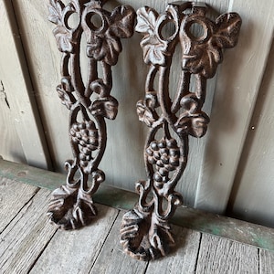 May include: Two cast iron decorative supports with a brown finish. Each support features a grape vine design with leaves and a cluster of grapes. The supports have a hole at the top and are designed to be mounted.
