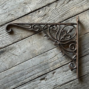 May include: A decorative, dark brown cast iron bracket with ornate scrollwork and a fleur-de-lis design. The bracket is L-shaped, with a curved end for hanging items. The metal has a rustic, aged appearance, suggesting outdoor use.