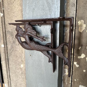 May include: A pair of cast iron shelf brackets with a mermaid design. The brackets are a dark brown color and feature a mermaid with flowing hair and a fishtail. The shelf brackets are mounted on a gray wooden surface.