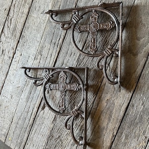 May include: Two ornate, cast iron shelf brackets. Each bracket features a decorative cross within a circular frame, with scrolling details. The brackets are finished in a dark brown color and are designed to be mounted on a wall.