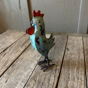 May include: A blue metal rooster figurine with a red comb and wattle. The rooster is standing on a wooden surface.