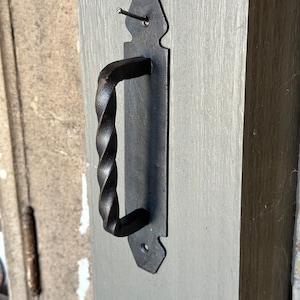 May include: Black metal door handle with a twisted design. The handle is attached to a rectangular plate with a decorative edge.