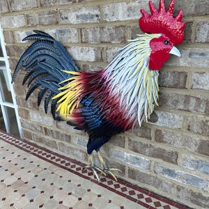 May include: A colorful metal rooster sculpture with red, yellow, blue, and white paint. The rooster is standing on a tiled floor with a brick wall in the background.