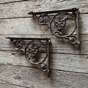 May include: Two antique cast iron shelf brackets. Each bracket features a decorative bird design within a circular frame. The brackets are dark brown with an aged, rustic appearance, and are mounted on a weathered wooden surface.