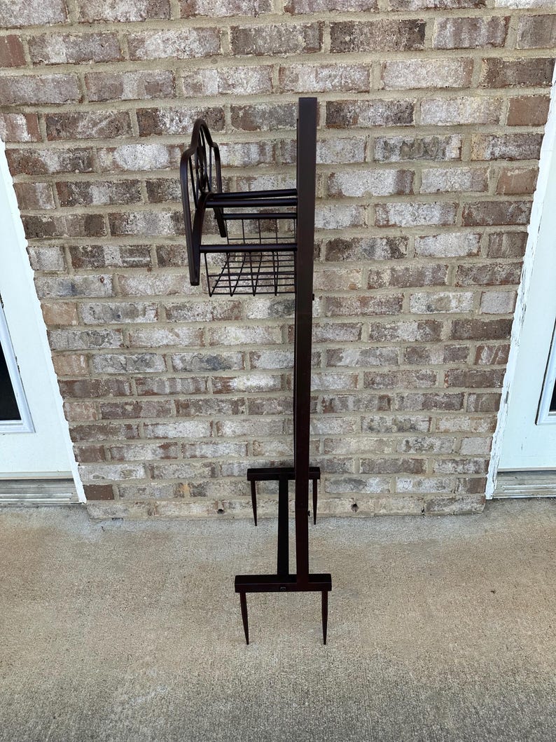 Op de afbeelding: Een donkerbruine metalen plantenstandaard met een draadmand en decoratieve rug. De standaard heeft een lange, verticale paal met een kleine plank en een mand. De basis heeft twee puntige poten.