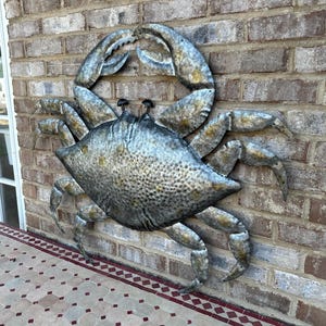 May include: A large, metallic crab sculpture mounted on a brick wall. The crab is silver with textured details and a weathered appearance. Its claws are raised, and it has small, dark eyes. The sculpture adds a coastal theme to the outdoor space.
