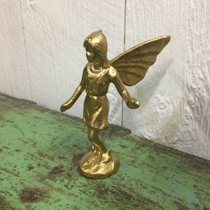 May include: A gold-painted metal figurine of a fairy with wings outstretched. The fairy is standing on a base and has a whimsical expression.