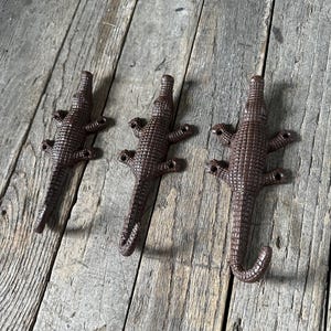 May include: Three brown metal wall hooks shaped like crocodiles. Each hook features a textured, scaly surface and four mounting holes located at the feet. The tail of each crocodile curves upward to form a functional hook for hanging items.