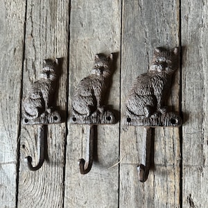 May include: Three cast iron cat hooks with a rustic brown finish. Each hook features a cat sitting with its paws tucked underneath. The hooks are mounted on a wooden surface.