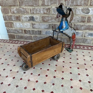 May include: A wooden wagon with metal wheels, pulled by a metal dog sculpture. The dog is made of recycled metal and painted in shades of blue, red, and white. The wagon is sitting on a tiled floor.
