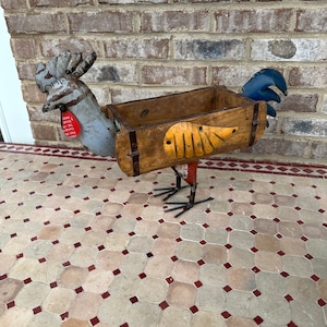 May include: A metal rooster planter with a wooden box for planting. The rooster is painted in shades of blue, gray, and red. The wooden box is painted brown with yellow accents. The rooster is standing on a tiled floor.