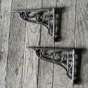 May include: Two cast iron shelf brackets with an ornate, scrolled design. The brackets are a dark gray color and are mounted on a weathered wooden surface. The brackets are designed to support a shelf.