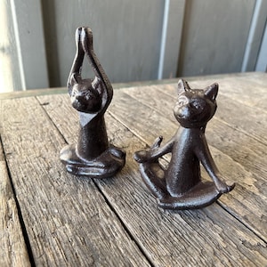 May include: Two dark brown cast iron cat figurines in yoga poses. One cat has its paws raised above its head, and the other is seated with its paws resting on its knees. The figurines are on a weathered wooden surface.