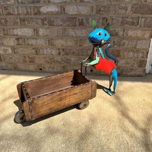 May include: A whimsical metal cat sculpture, painted blue with green ears and a red outfit, pulls a wooden wagon. The wagon is brown with metal accents and wheels. The sculpture is outdoors, in front of a brick wall.
