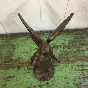 May include: A cast iron angel figurine with outstretched wings. The angel is standing on a round base and is painted in a dark brown color.