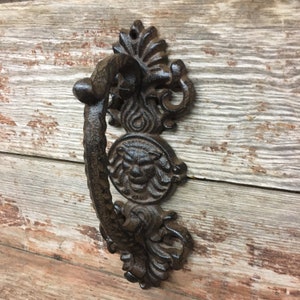 May include: A cast iron door knocker with a lion's head design and ornate scrollwork. The knocker is dark brown and has a rustic finish.