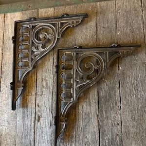 May include: Two decorative, cast iron shelf brackets with an ornate, scroll design. The brackets are dark gray with black accents and are mounted on a weathered, wooden surface. The brackets are designed to support a shelf.
