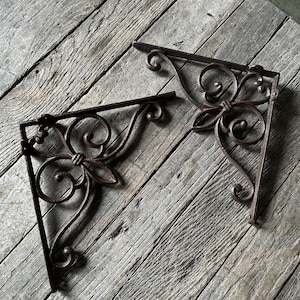 May include: Two ornate, dark brown metal shelf brackets with a floral and scroll design. The brackets are angled and appear to be vintage or antique, with a rustic finish. They are set against a weathered, gray wooden surface.