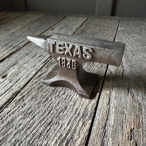 May include: A cast iron anvil with the word "TEXAS" and the year "1836" embossed on the top. The anvil is a dark, metallic color and sits on a weathered wooden surface.