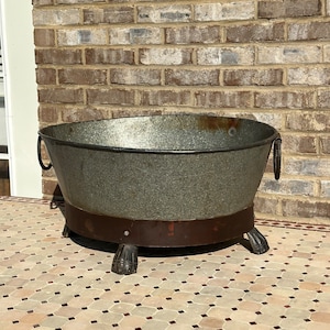 May include: A large, round, galvanized metal planter with a brown metal base and four decorative feet. The planter is sitting on a tiled surface.