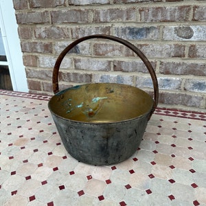 May include: A vintage brass pot with a worn black finish and a rusty iron handle. The pot is sitting on a tiled surface.