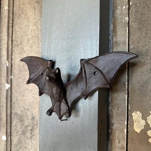 May include: A dark brown, cast iron bat-shaped wall hook with outstretched wings. The bat features a textured surface and two small circular holes in the wings for mounting purposes, securely attached to a vertical gray wooden board surface.