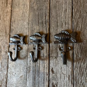 May include: Three cast iron fish-shaped wall hooks. Each hook features a detailed fish design above a curved hook. The hooks are mounted on a wooden surface, showcasing a rustic aesthetic. The hooks are ideal for hanging coats, hats, or other items.