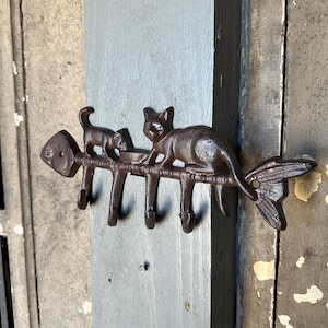 May include: A cast iron key holder shaped like a fish with three cats on top. The cats are in various poses, including one holding a bowl. The key holder has five hooks for hanging keys.