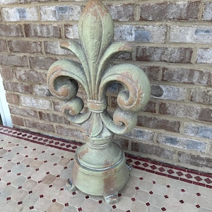 May include: A weathered, sage green fleur-de-lis sculpture with a pedestal base. The decorative piece has a distressed finish with brown accents. The sculpture is placed on a patterned tile surface in front of a brick wall.