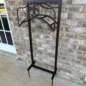 May include: A dark brown metal garden hose holder with a fleur-de-lis design. The holder has a rectangular frame, a curved top, and a wire basket for storage. The holder is designed to stand upright.